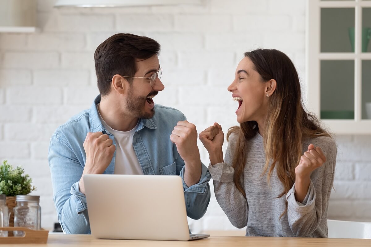Un couple devant PC se regarde, poings serrés en signe de victoire, hyper heureux de leur RED Box Fibre à moins de 21€ avec frais de mise en service offerts